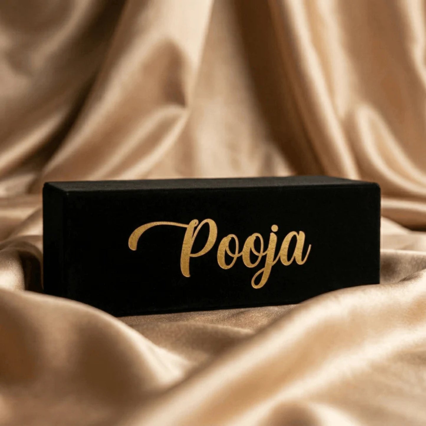 Black rectangular box with 'Pooja' written in gold on a beige fabric background
