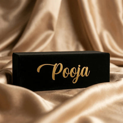 Black rectangular box with 'Pooja' written in gold on a beige fabric background